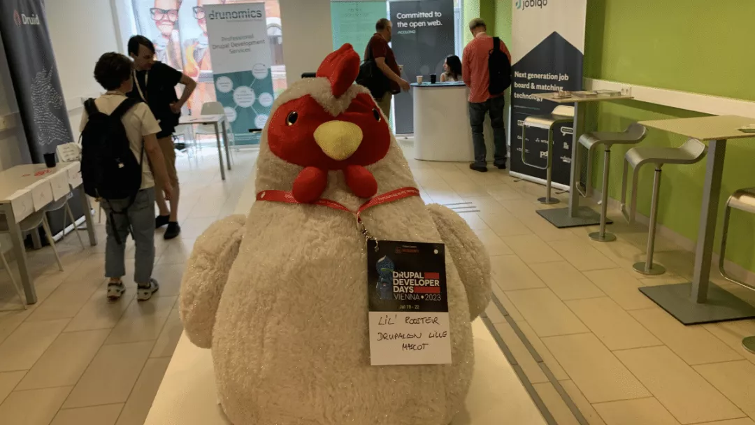 A plushed chicken is sitting in front of the camera, with a Drupal Dev Days 2023-Badge. He obviously is an attendee. His badge says "Lil' Rooster, Drupal Con Lille Mascot.
